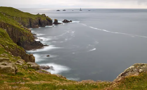 This is a landscape photograph taken on a spring evening, capturing the rugged cliffs and sweeping grassy coastline of Cornwall. The image prominently features the sea with a smooth, misty appearance achieved through long exposure, accentuating the motion of the waves against the rocks. In the distance, the Longships lighthouse rises above the surrounding sea and rocky outcrops, serving as a notable landmark. The cliffs, covered in green vegetation, dominate the foreground, framing the expansive view towards the Longships lighthouse and the surrounding waters.
